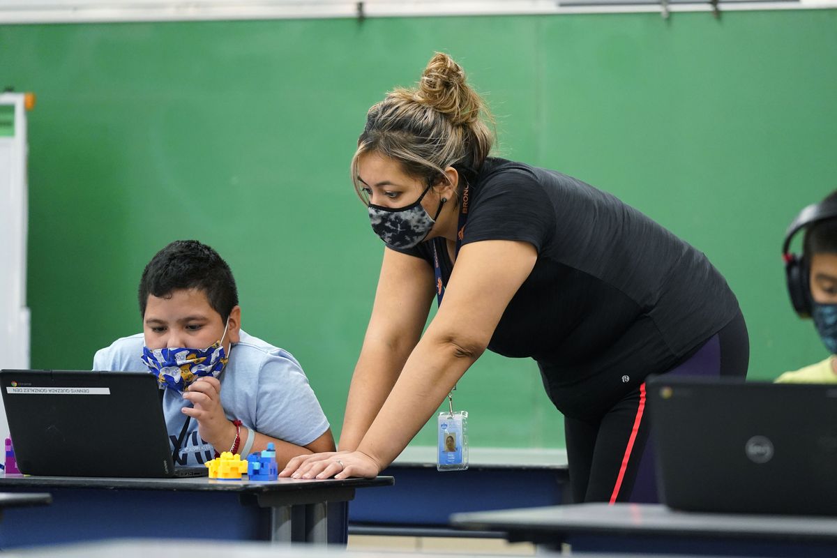 Masked teacher helps masked students who are working on laptops.
