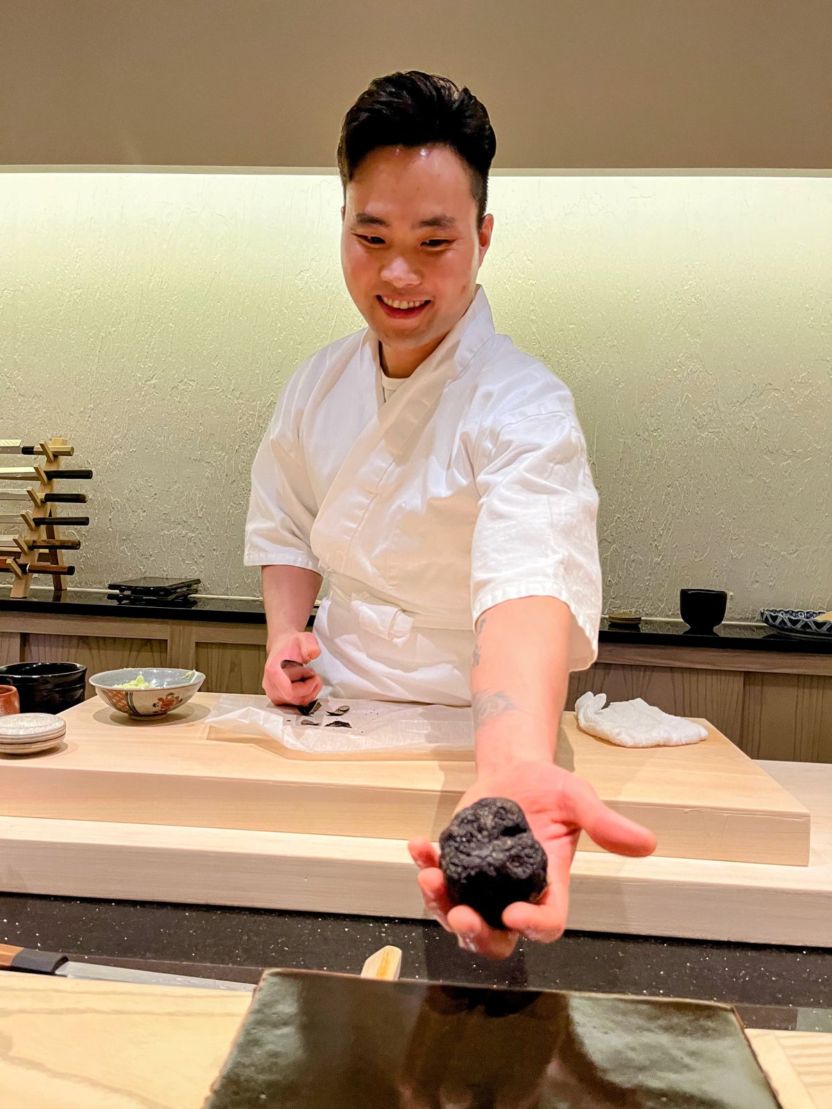 A man in a white chef’s coat stands behind a sushi counter with one hand extended, showing off a large black truffle.