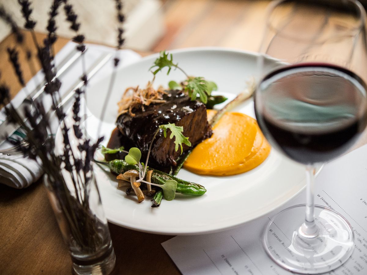 A glass of red wine and flowers in front of a white plate with a saucy meat dish at Tuome.