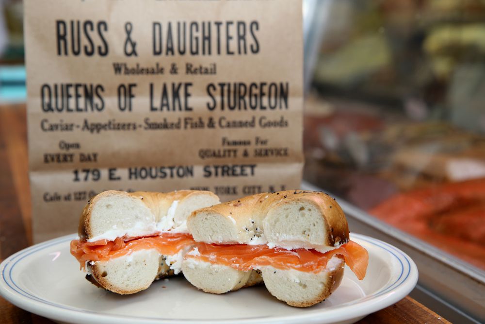 Pieces of salmon jut out of a bagel sandwich sliced in half, that’s placed on a white cermaic plate. A sign for Russ &amp; Daughters hangs in the background.
