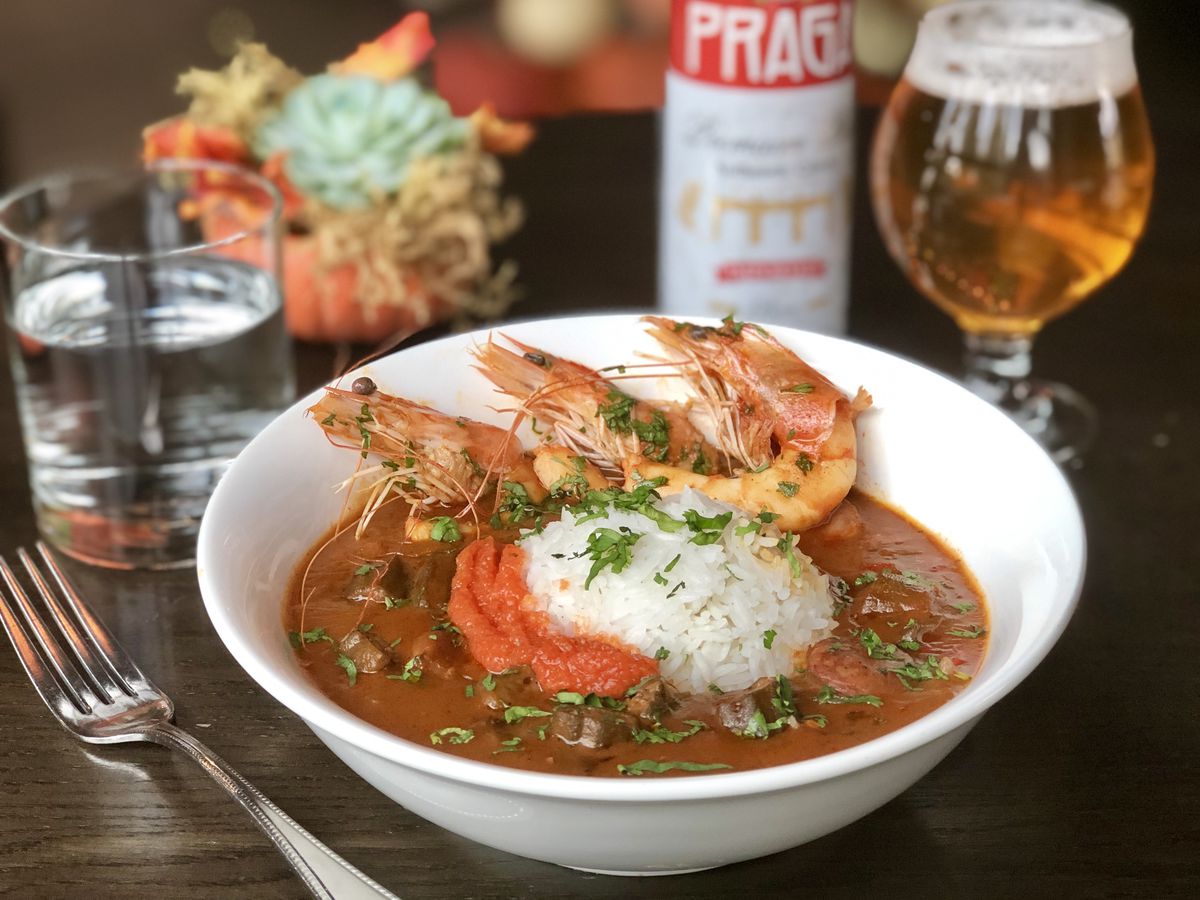 A bowl of shrimp gumbo with rice and a glass of beer in the background.