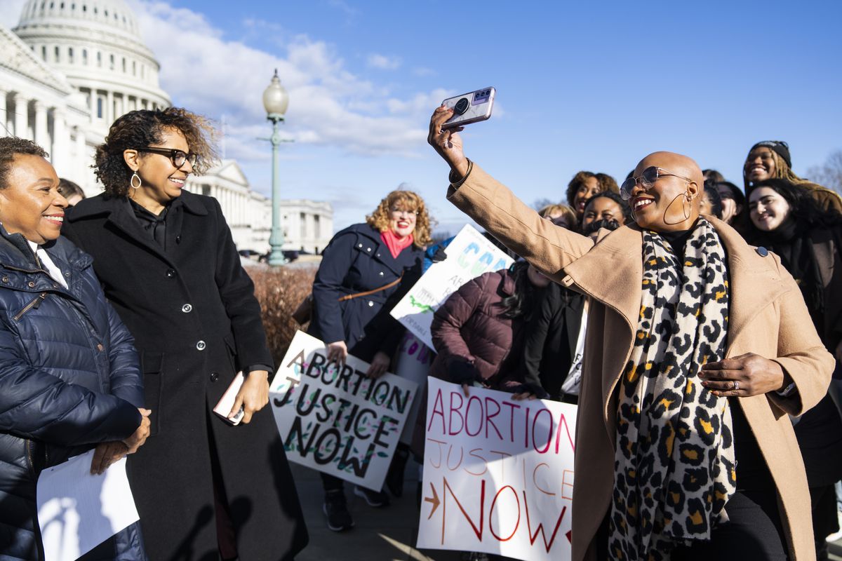 Pressley, her head shaved and in a camel overcoat, smiles as she poses with women holding signs reading “Abortion justice now.” The Capitol dome looms in the background.