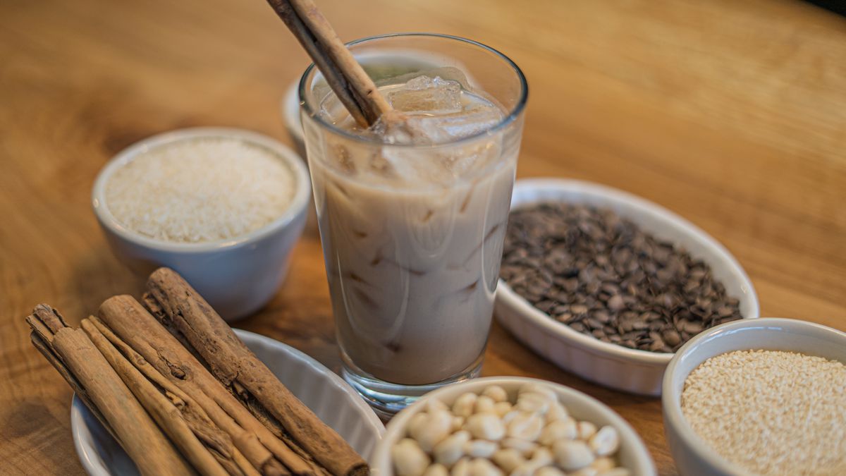 A glass pint glass filled with a brown beverage and ice surrounded by little white bowls filled with rice, sesame seeds, sunflower seeds, and cinnamon sticks.