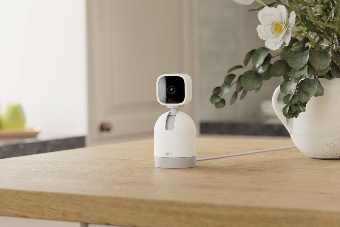 The scene is a wooden kitchen counter with a flower pot offset to the right, and in the center is the Blink Mini cam, which is a blocky black and white device. It’s mounted on a white bullet-shaped body that makes it look like a Pixar-style robot device. There’s a cable running out the back.