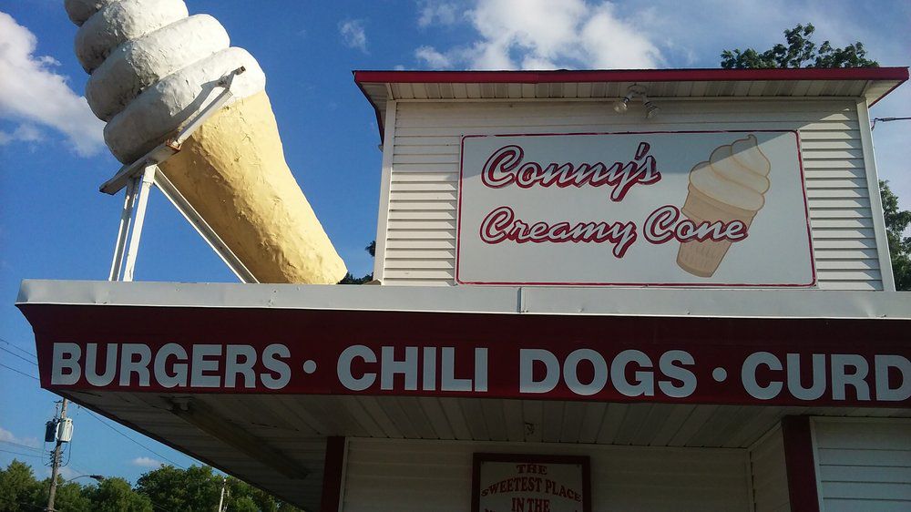 The exterior of a building that says “Conny’s Creamy Cone” and has an awning reading “burgers chili dogs) and a large sculpture of an ice cream cone. 