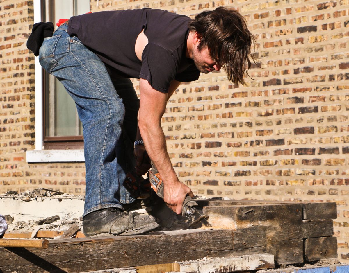 A white man stands on old dusty wood in jeans and a black t-shirt bending over with a power saw to cut a sample of dark wood.