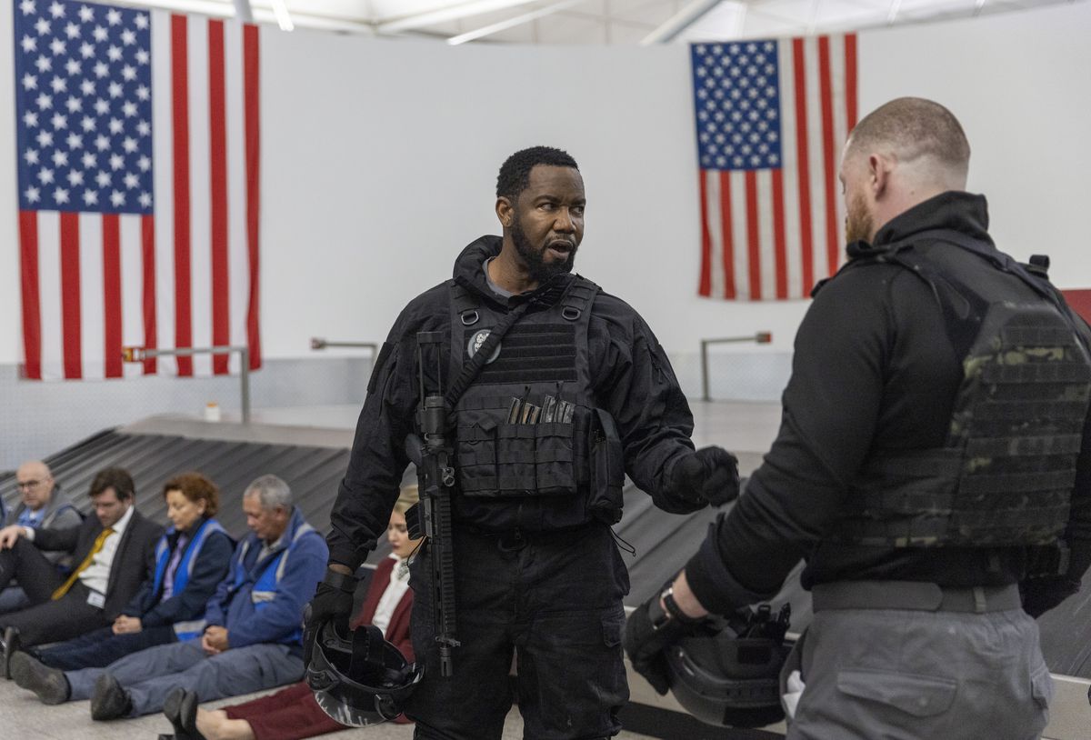Michael Jai White, wearing a bulletproof vest and with a rifle hanging on his shoulder, talks to another man wearing a bulletproof vest while hostages are lined up against the airport baggage carousel in One More Shot.