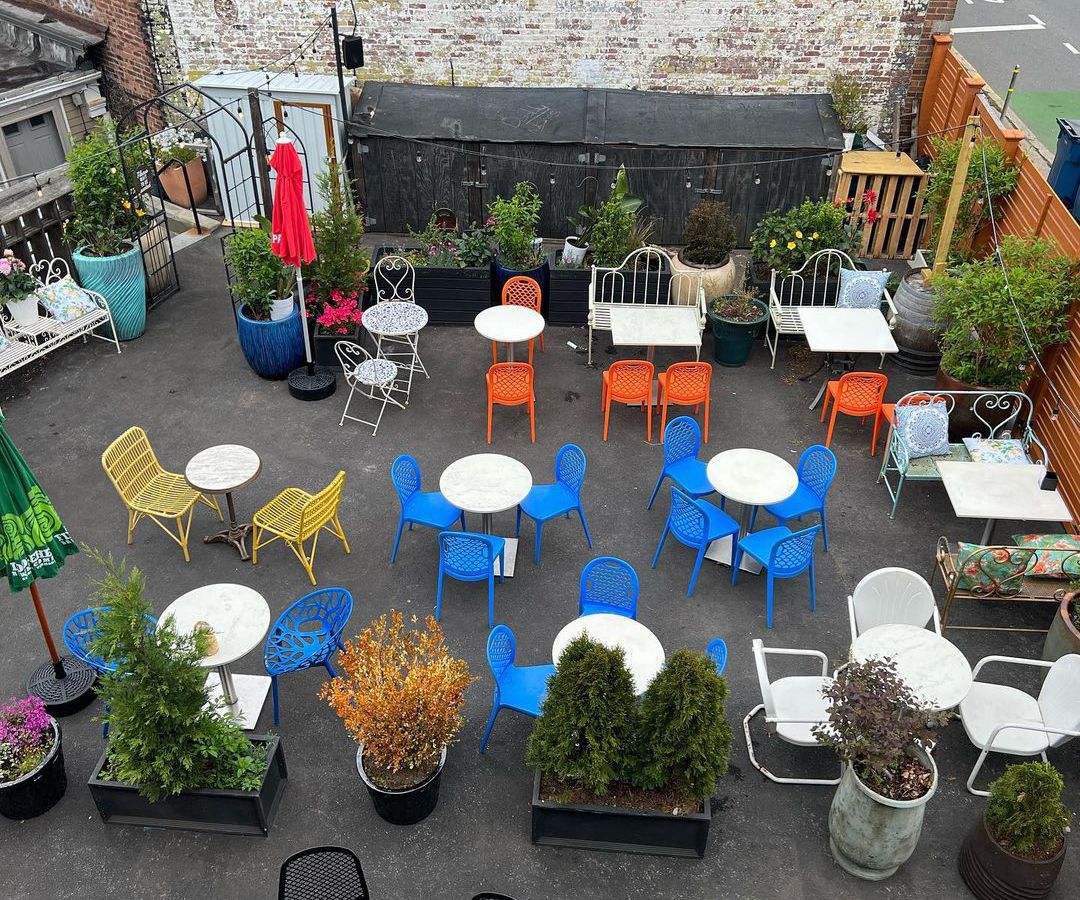 Overhead view of a restaurant patio with an assortment of colorful metal chairs, round white tables, trees, and umbrellas.