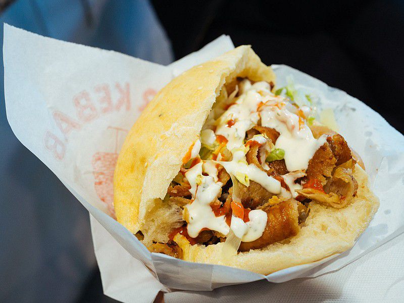 A pita split open into a sandwich stuffed with kebab meat and diced vegetables, covered in white sauce, all held in wax paper.