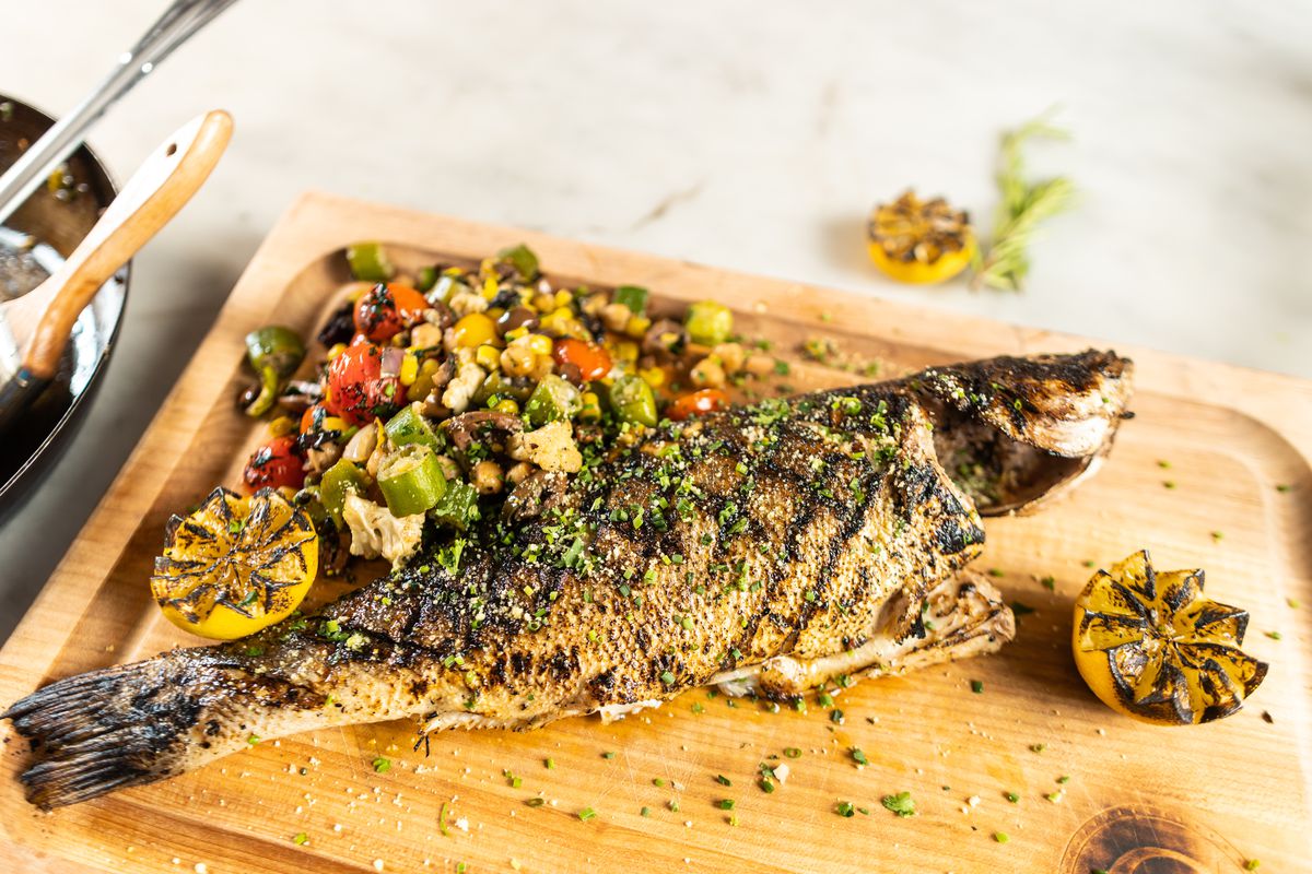 A whole roasted branzino rests on a wooden board. It is garnished with chopped fresh vegetables and two roasted lemon halves, plus spices that decorate the board.