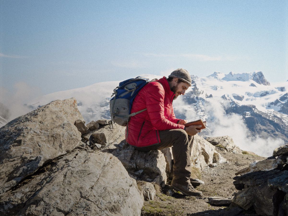 Pietro (Luca Marinelli) in hiking gear sits atop a mountain reading a book, with more snow-topped mountains behind him, in The Eight Mountains