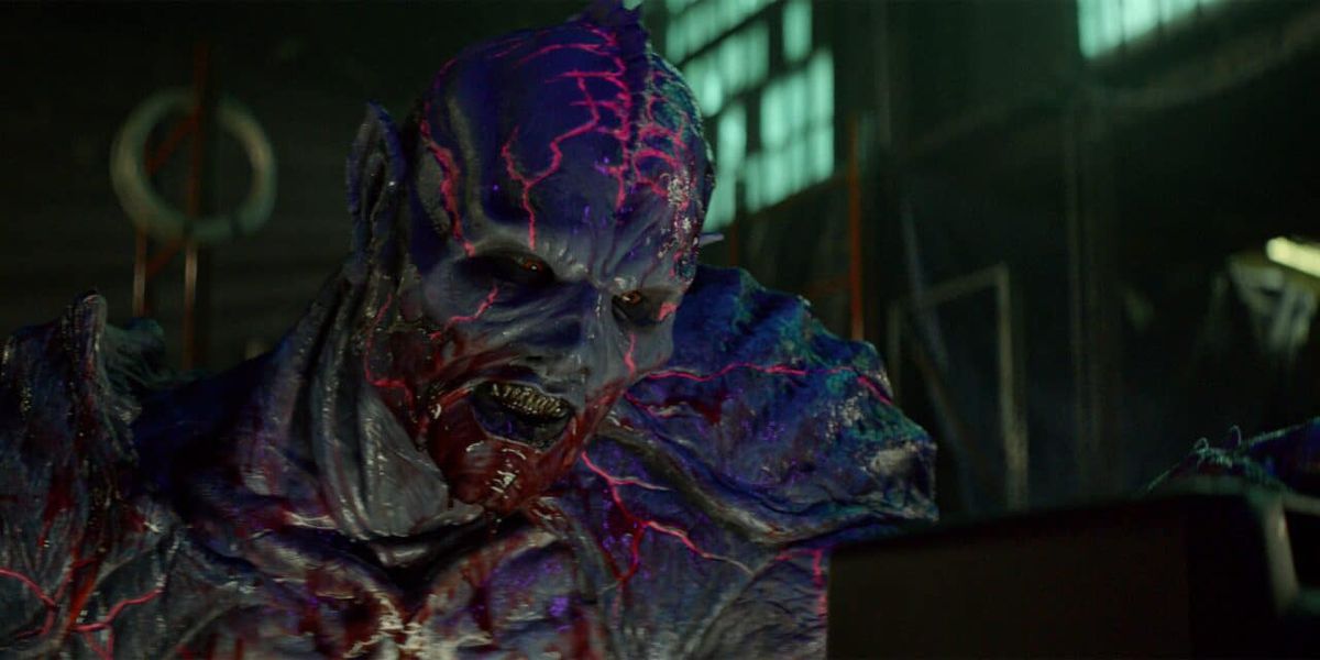 A purple, reptilian alien creature with human features, red eyes, glowing pink veins, and fang-like teeth leers ominously at something off-screen.