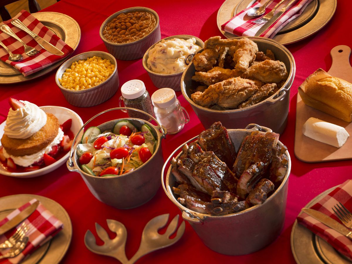 Buckets of fried chicken, salad, dessert, and sides like mac and cheese and beans, on a red table cloth nearby plates set for dinner.