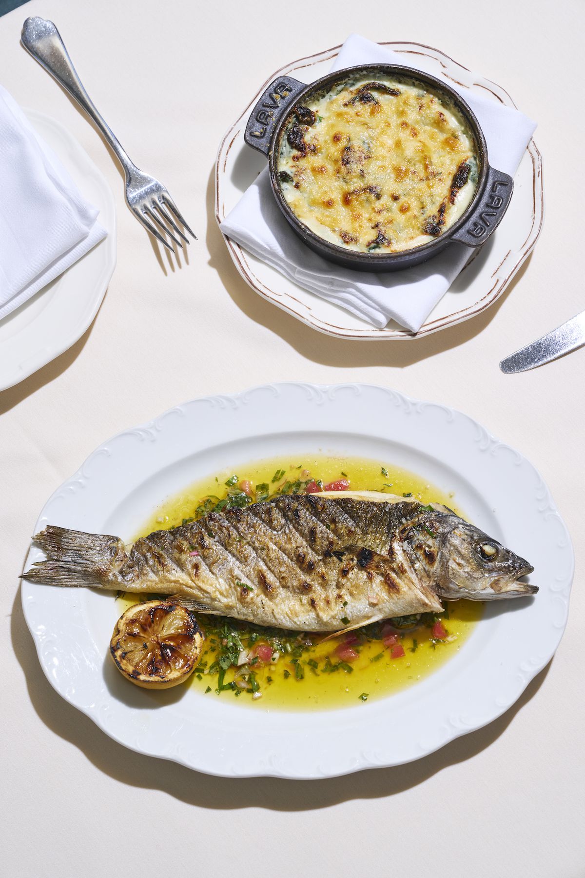 A horizontal grilled fish with head on, on a scalloped plate with side dish, against a bright white table at new restaurant Shirley Brasserie.