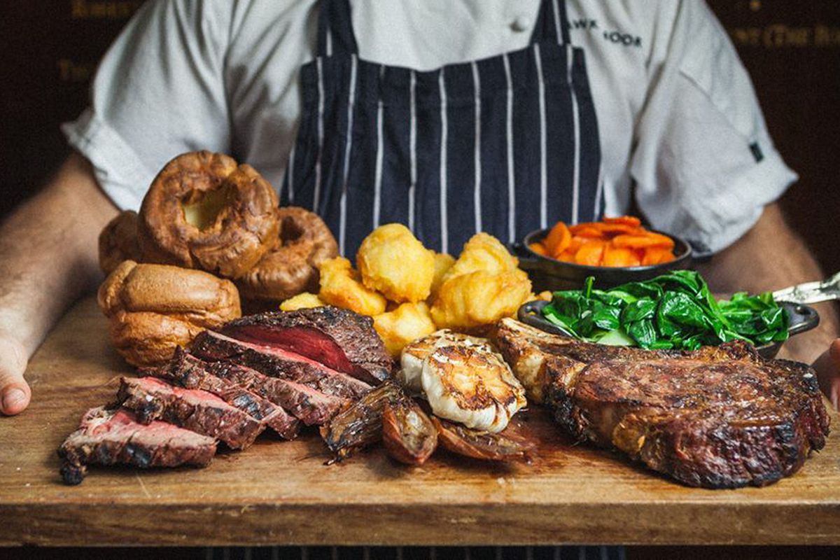 A board of beef, potatoes, and Yorkshires from Hawksmoor’s U.K. operation, held by a chef in a classic navy-and-white-striped apron