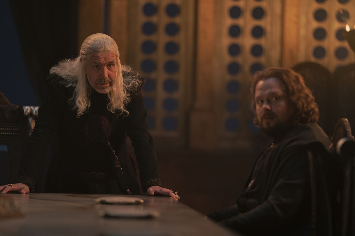 King Viserys and Lord Strong at the table of the Little Council;  Viserys stands and looks up while Viserys sits and turns to look the same 