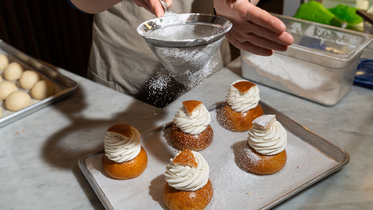 A baker dusts powdered sugar over a baking sheet of five Semla pastries 