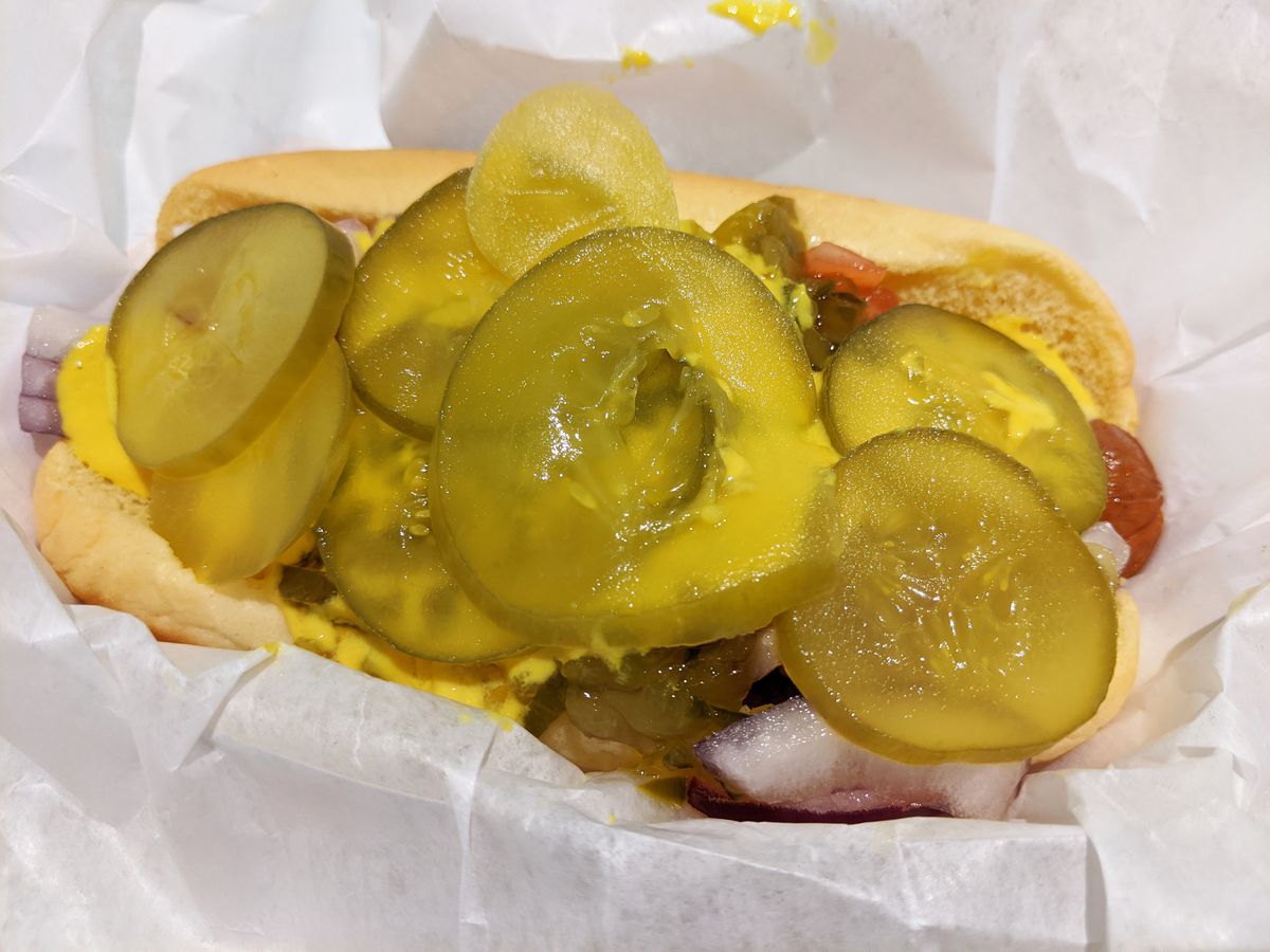 A hot dog in a plain bun is concealed under a pile of pickle chips and watery floe of yellow mustard.