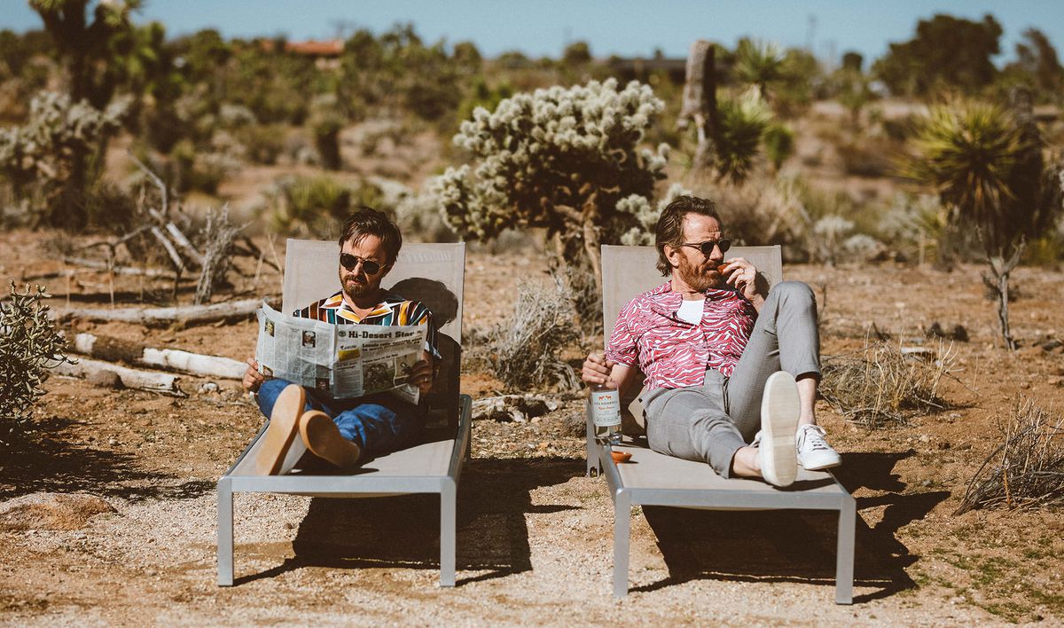 Two bearded dudes lay on lounge chairs in the desert. One reads a newspaper and the other oh-so-casually grasps a bottle of liquor.