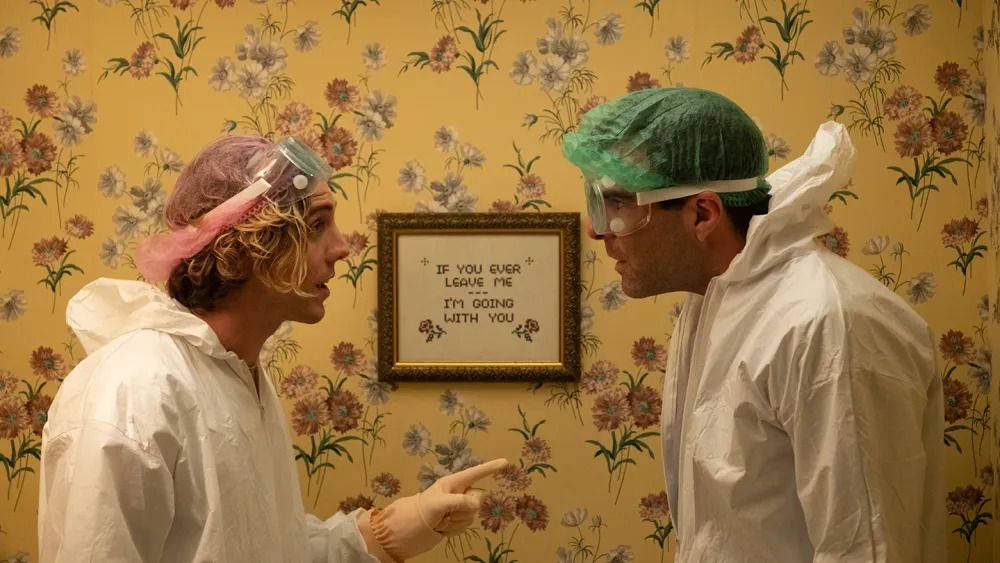 Two men wearing white hazmat suits and goggles stand in front of a wall decorated in yellow floral wallpaper.