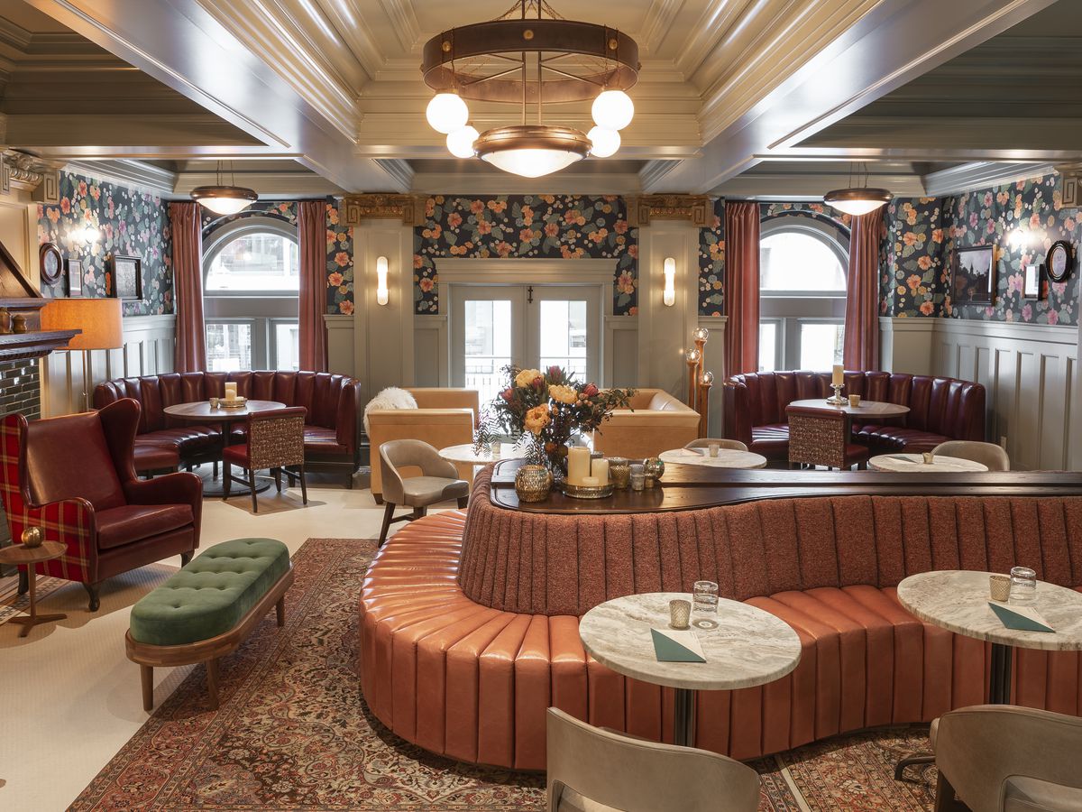 Pink swirling couches fill a hotel bar with floral wallpaper and an unlit fireplace