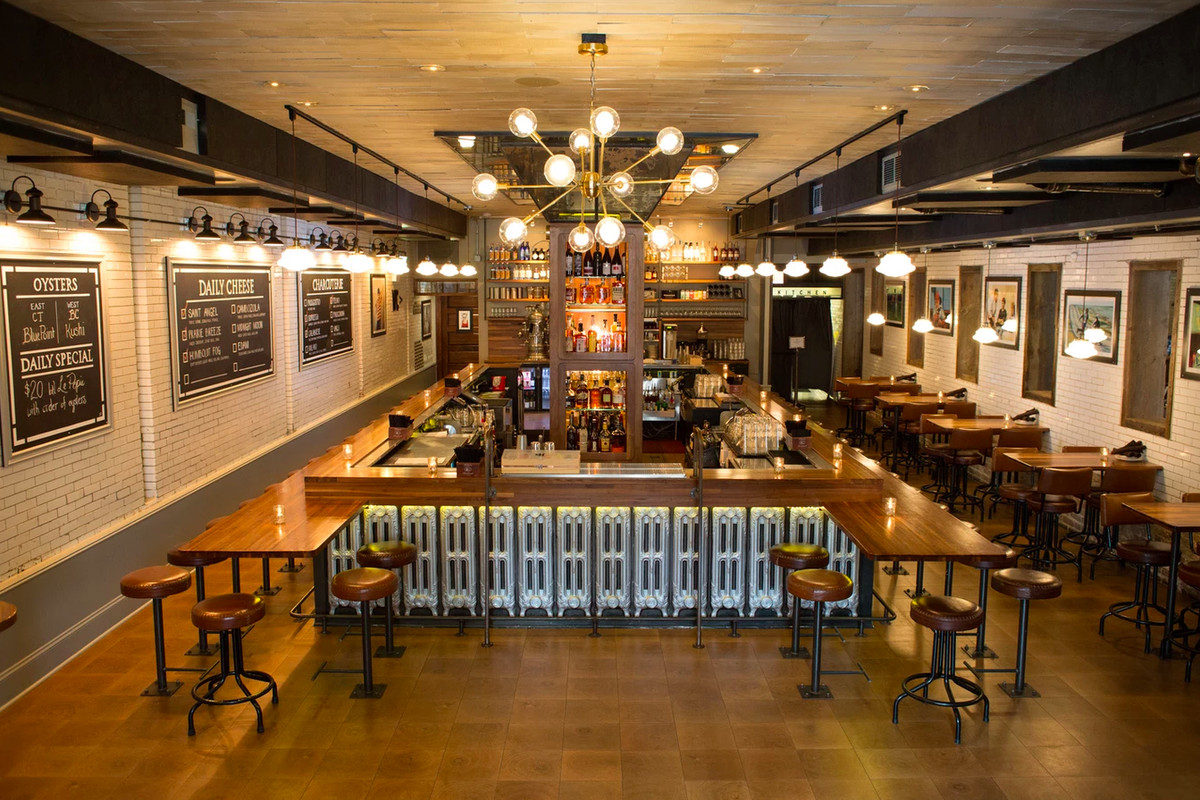 A view of high above a sunken bar room with a large rectangular central wooden bar, wooden tables, brass fixtures, twinkling lights, and chalkboards on the white brick walls.