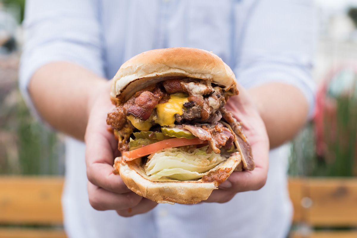 A tall meaty burger with cheese and lettuce and pastrami and chili, in hands.