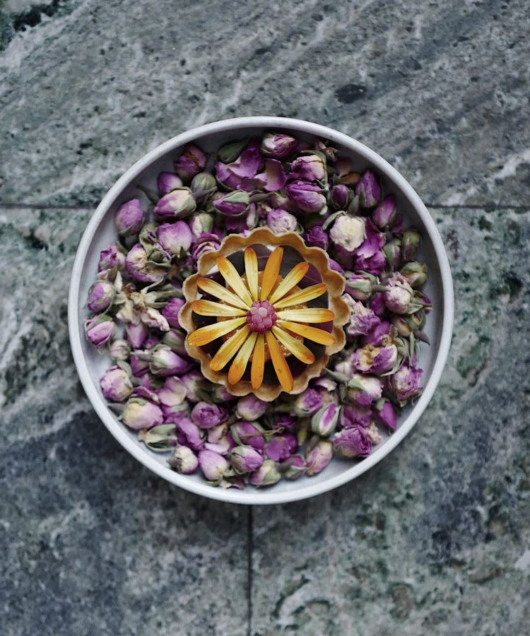 From above, yellow flower petals cover a dish set in a larger bowl of flower buds on a marble background