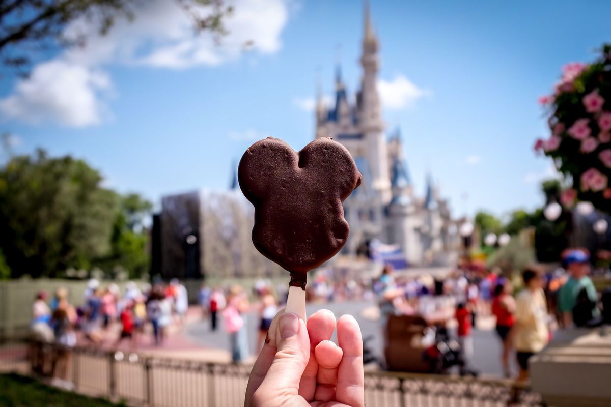 A hand holds a chocolate treat shaped like Mickey Mouse’s head in front of the Magic Castle.