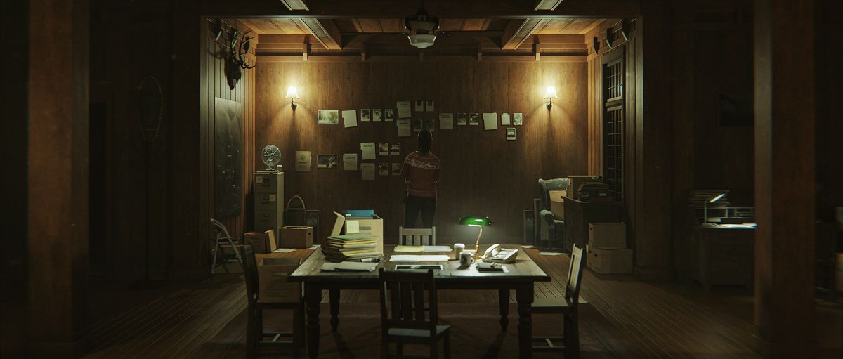 An image of Saga standing in an office room. It’s dimly lit and there is a desk filled with papers and a wall with papers taped up in Alan Wake 2