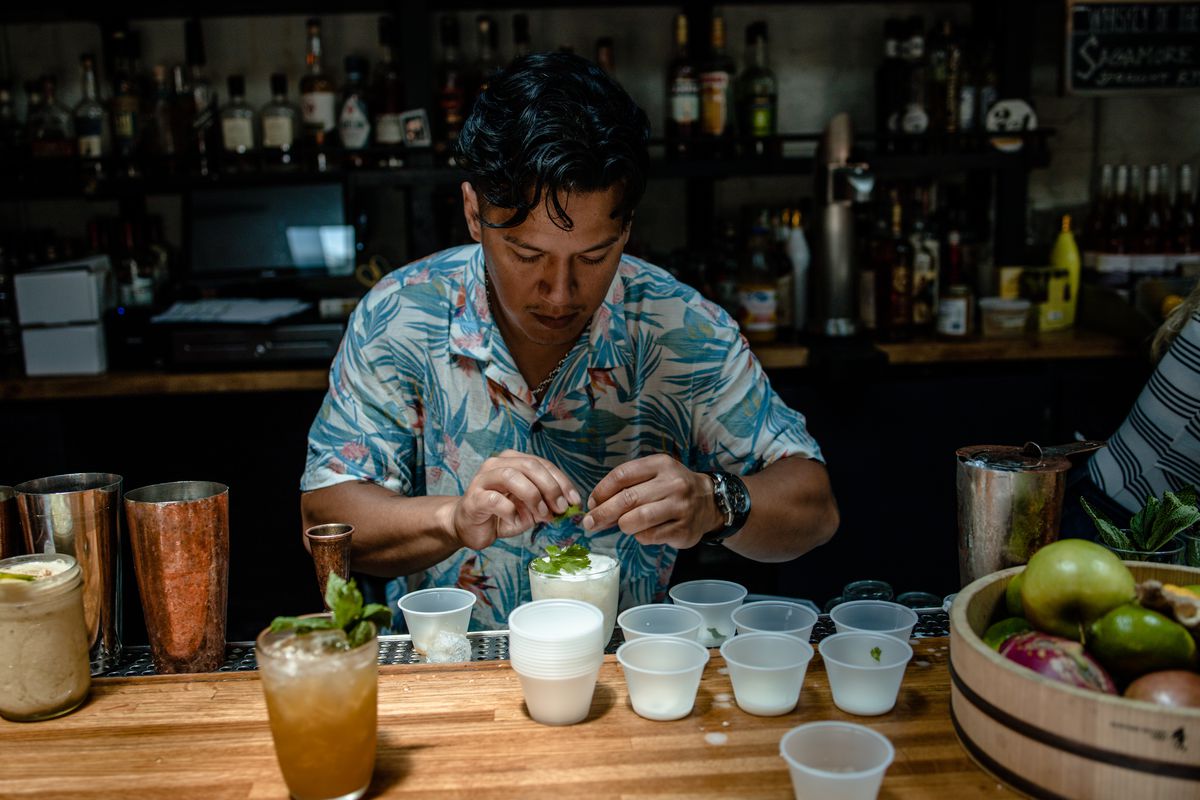 A man in a tropical-print shirt makes a frothy white drink, holding leafy garnish, as he stands behind a dimly lit bar. 