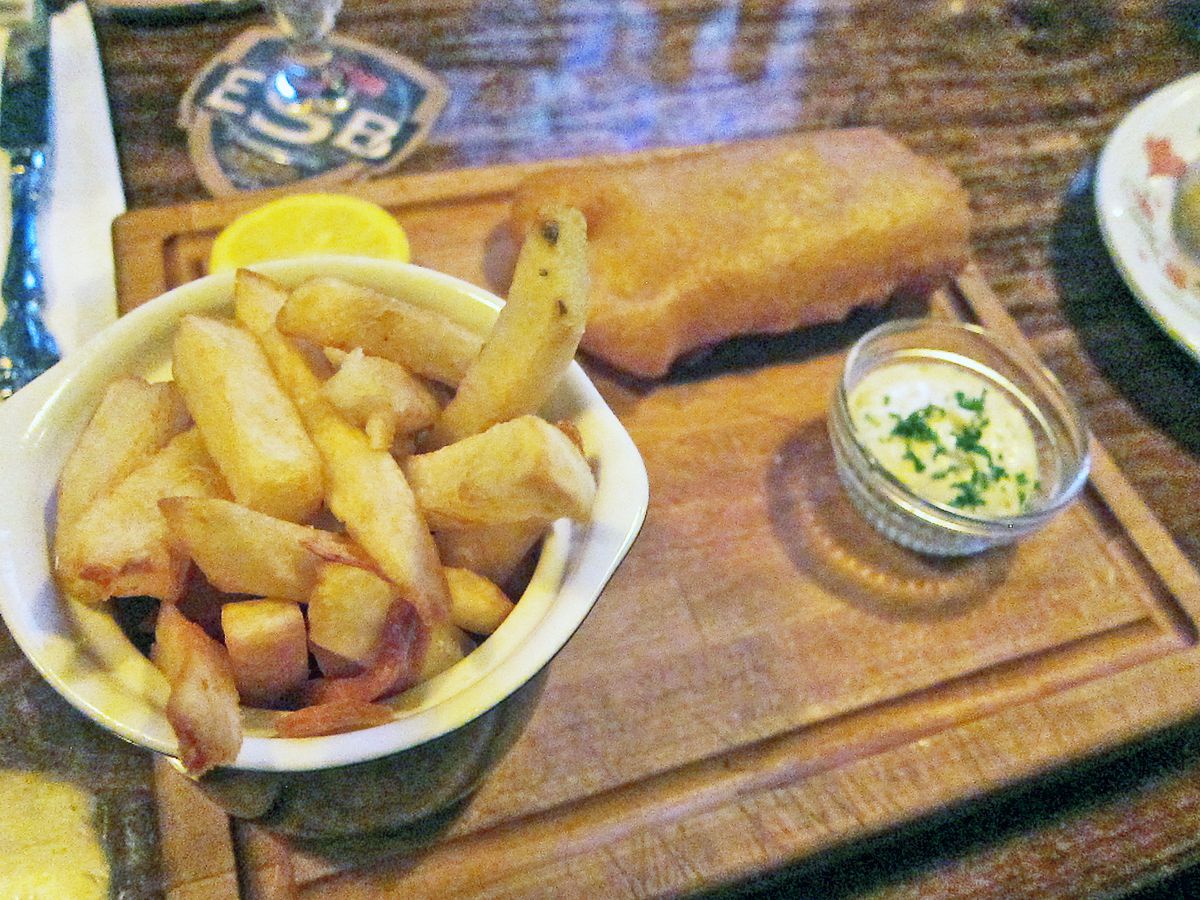 A breaded fish filet on a wooden cutting board and chips in a bowl.
