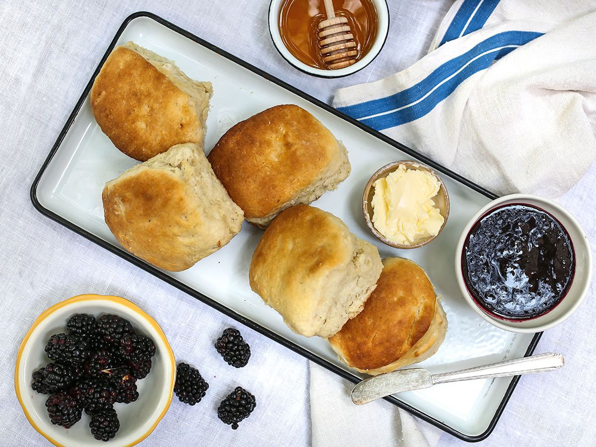 Bird’s-eye view of a white table holding a rectangular white platter full of biscuits, butter, and blackberry jam. Smaller ramekins of fresh blackberries and honey flank the biscuit tray.