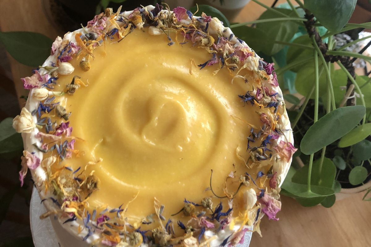 A hand holds a white cake covered in edible flowers, with a swirl of golden yellow passionfruit curd on top from Bee’s Cakes.