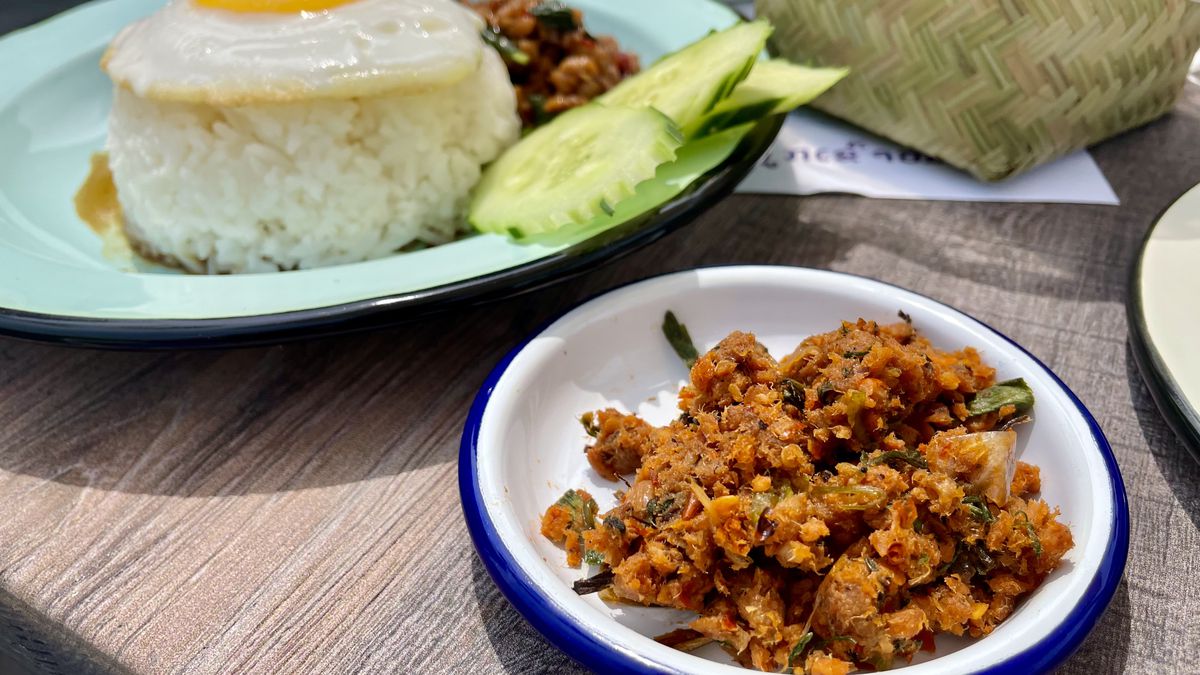 A small white dish of jeow (an orange, spicy, salsa-like Lao dipping sauce) on a table with a larger blue plate of rice, eggs, cucumber, and chicken in the background. 