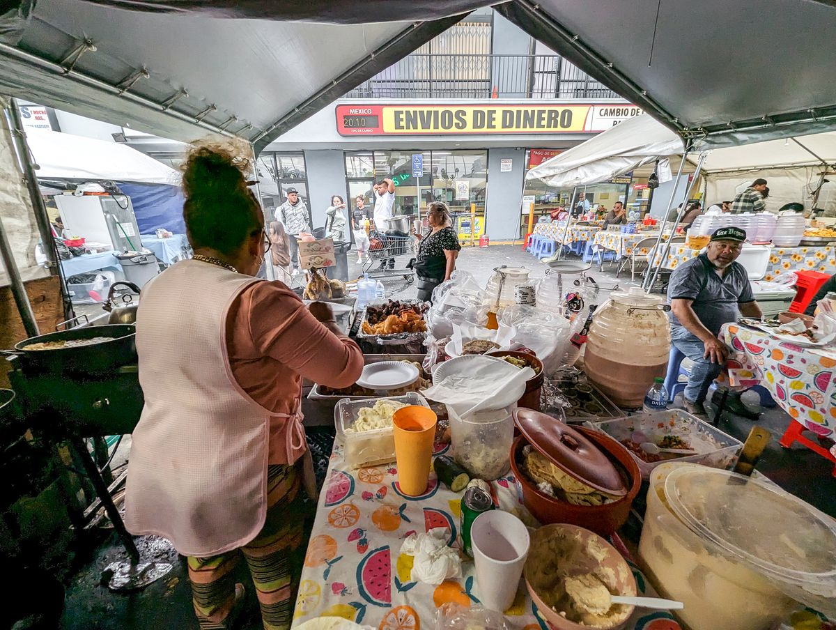 A female vendor with a white apron and apricot colored shirt looks away from camera as she prepares food from various tubs at a daytime street food vendor market in Los Angeles.