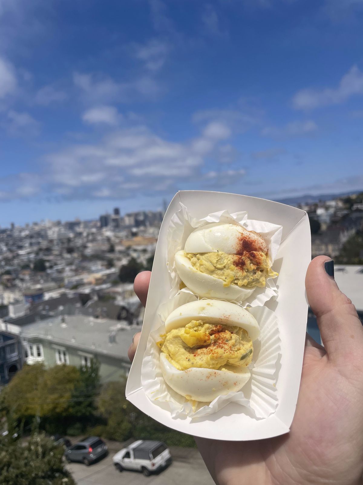Two deviled eggs in a paper tray are seen against a city background.
