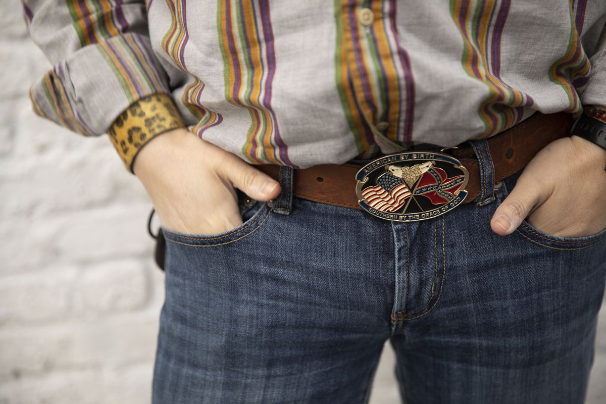 Closeup of waist and thighs with button-down tucked in and a metal belt buckle depicting the American and Confederate flags.
