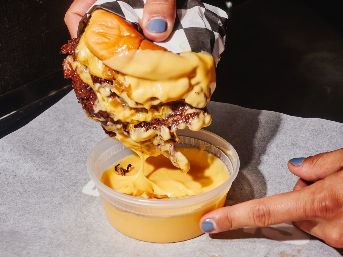A hand with painted nails dips a triple smash burger on a potato roll into a plastic tub of neon yellow cheese.