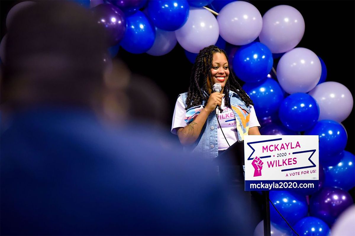 Democratic candidate McKayla Wilkes onstage holding a microphone and smiling while standing under a balloon arch.