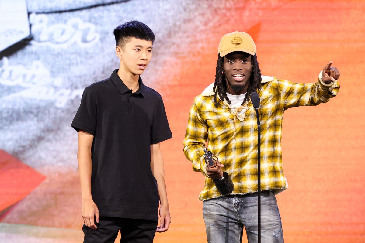 A photo of Kai Cenat on stage at the 2-23 Streamy Awards. He standing next to another creator and is holding an award in his hand as he talks into the mic. 