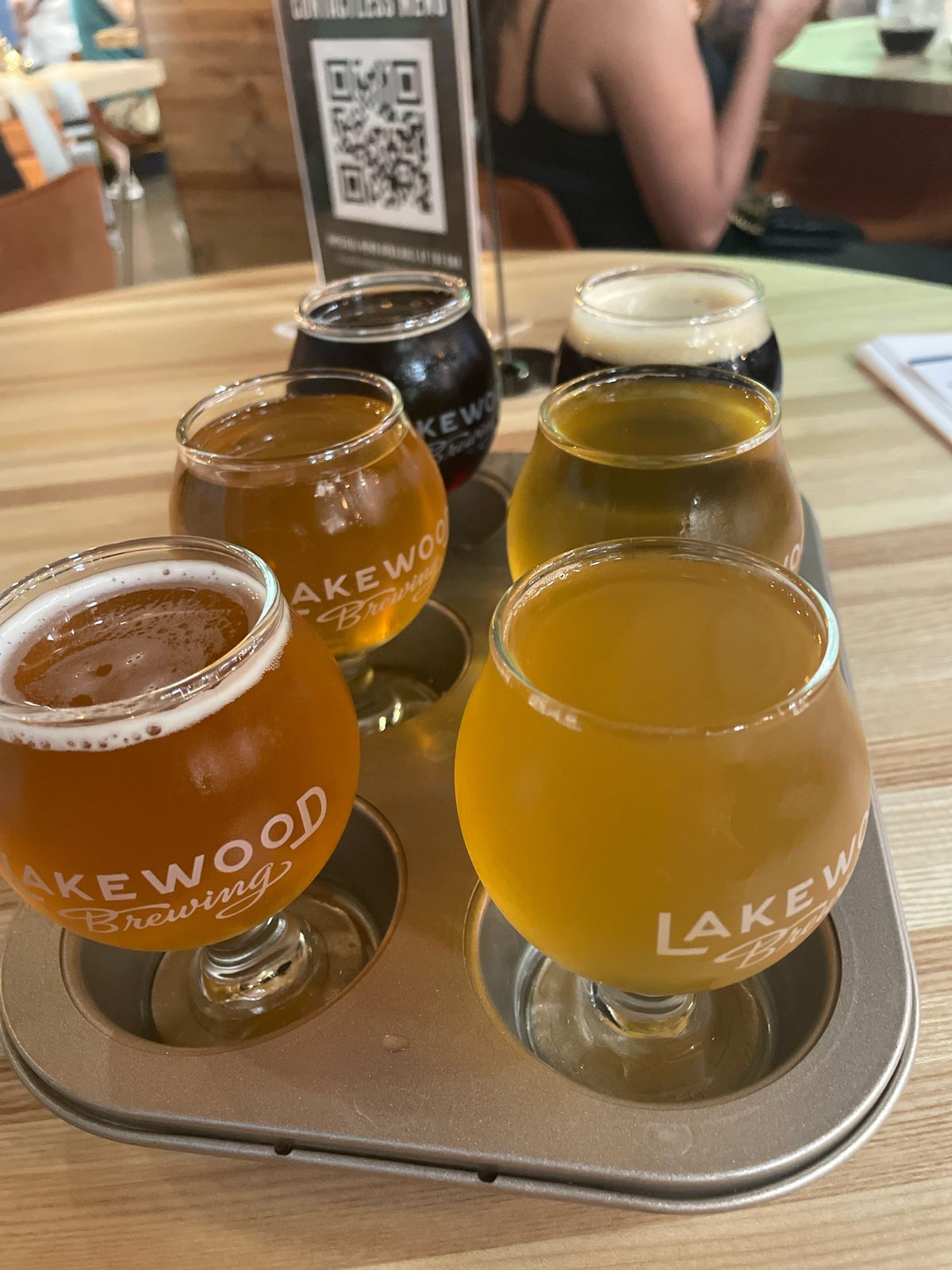 In a muffin tin, a collection of tasting sized glasses are filled with various beers. The glasses are emblazoned with “Lakewood Brewing” in white letters.