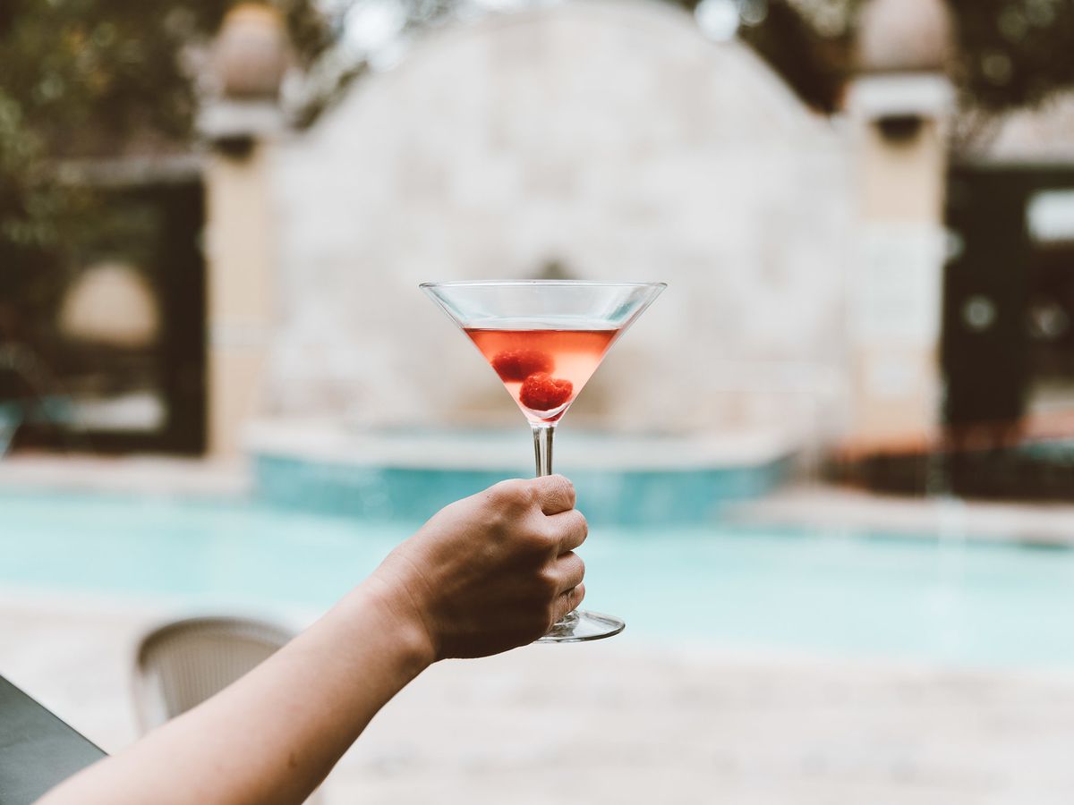 A woman’s hand holds up a cherry martini in front of a small pool.