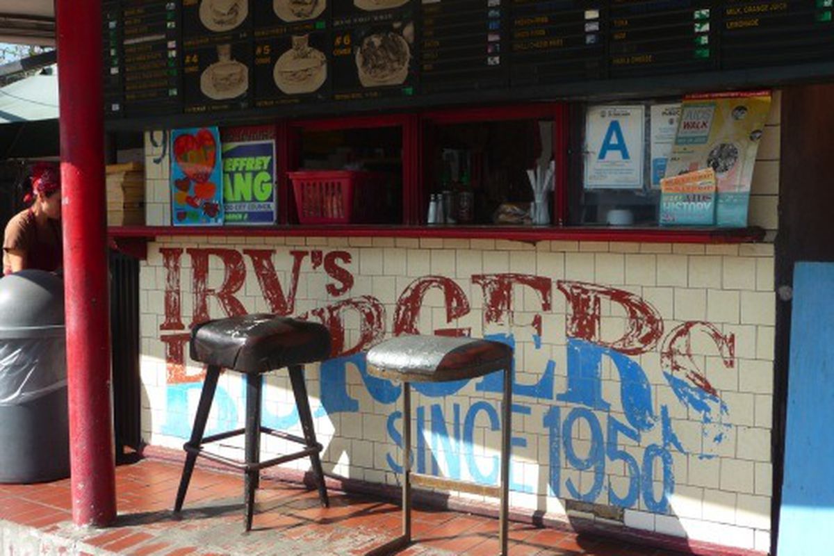 A historic and well worn burger stand with askew stools and faded signage, in daytime.
