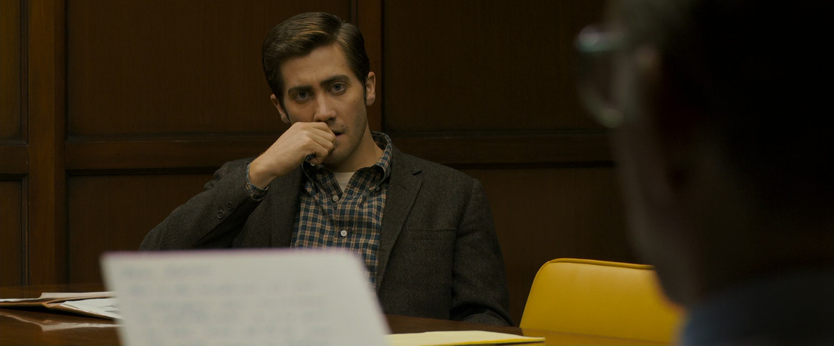 Jake Gyllenhaal, wearing a suit and a button-up collared shirt, bites his nails while looking at a man reading a piece of paper in Zodiac