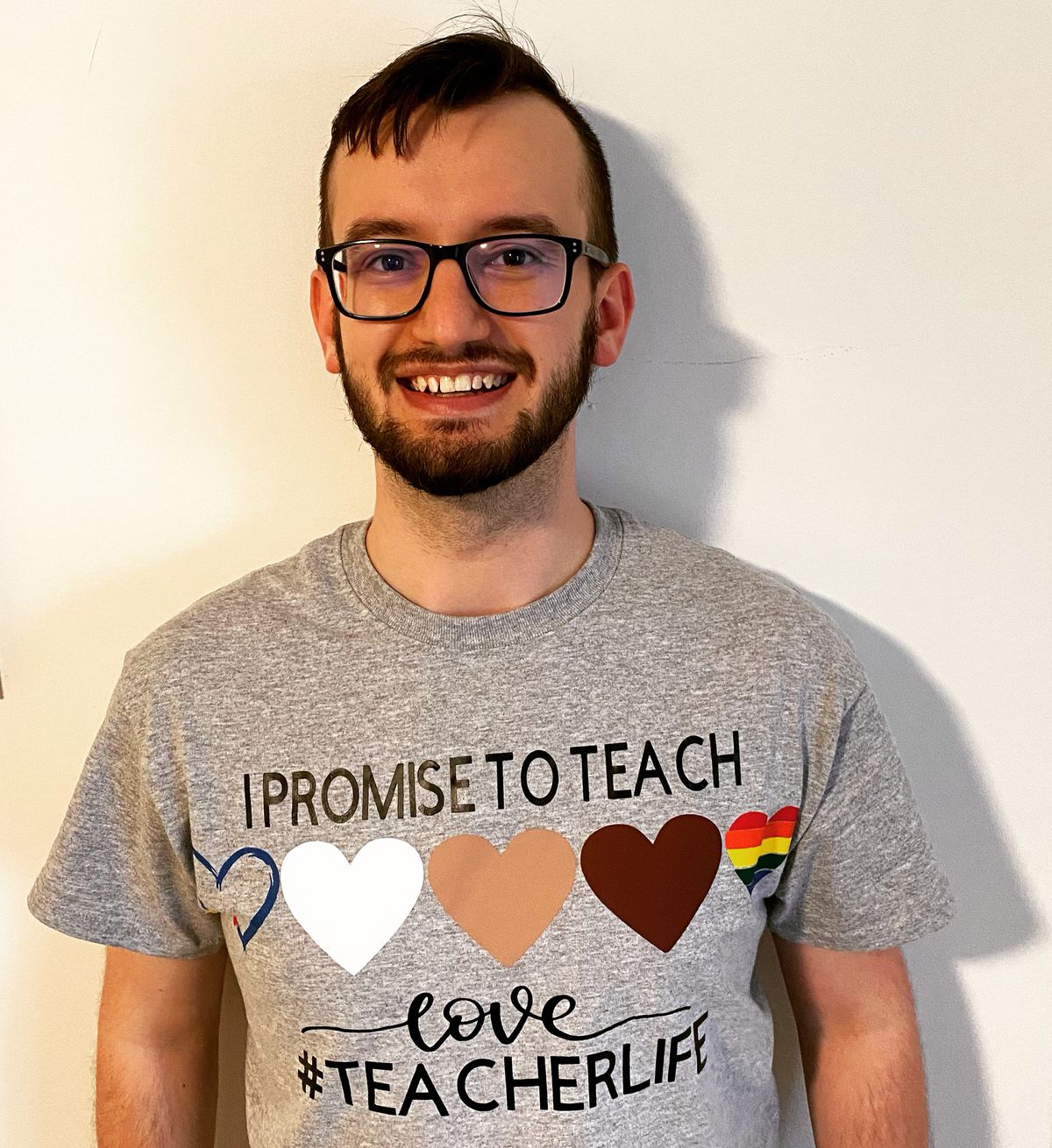 Man in gray shirt bearing hearts of different colors. Shirt says, “I promise to teach love #teacherlife”