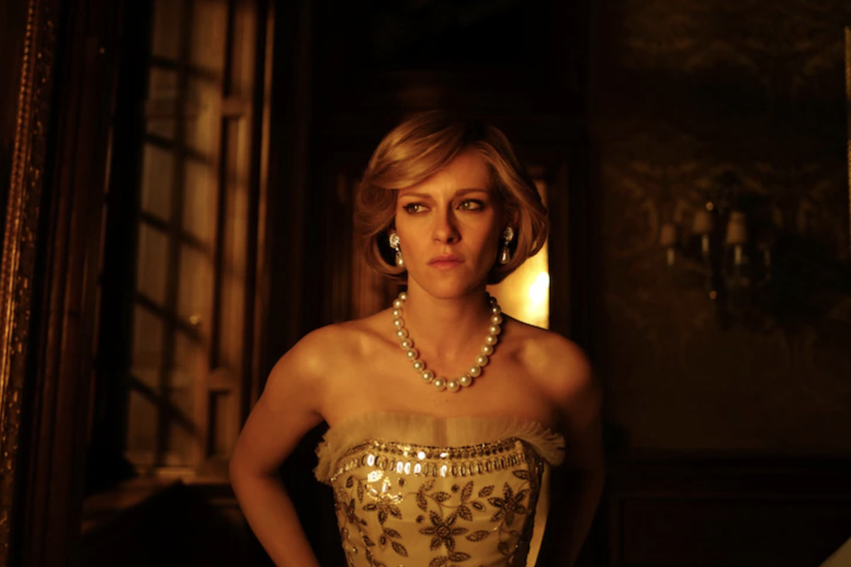 Kristen Stewart as Princess, standing in the darkened hallway of a palace wearing a strapless gold evening gown and a string of pearls, in Pablo Larraín’s “Spencer.”