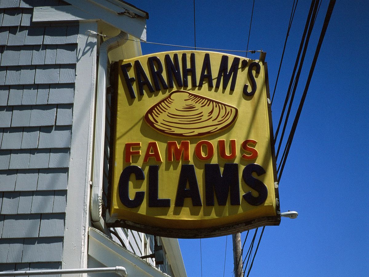 Closeup on the yellow rectangular sign on the side of a gray-shingled restaurant exterior. The sign reads “Farnham’s Famous Clams” and includes a clam.