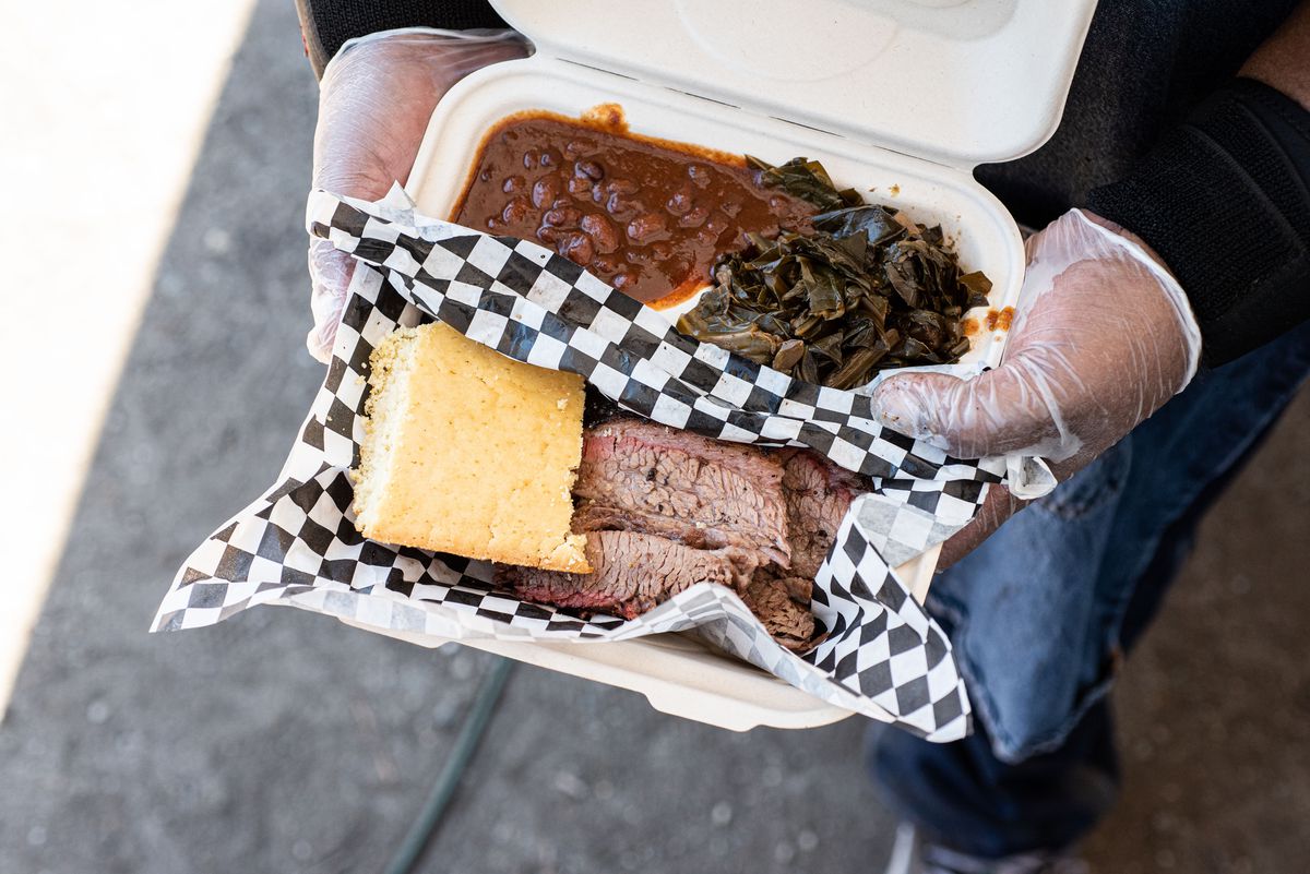 An overhead styrofoam plate of brisket, cornbread, beans, and greens from a barbecue restaurant.