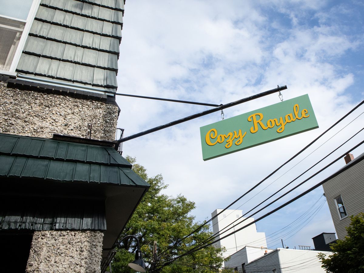 A hand-painted yellow and light green sign hangs about a restaurant with the words “Cozy Royale.” The sky is blue with clouds.